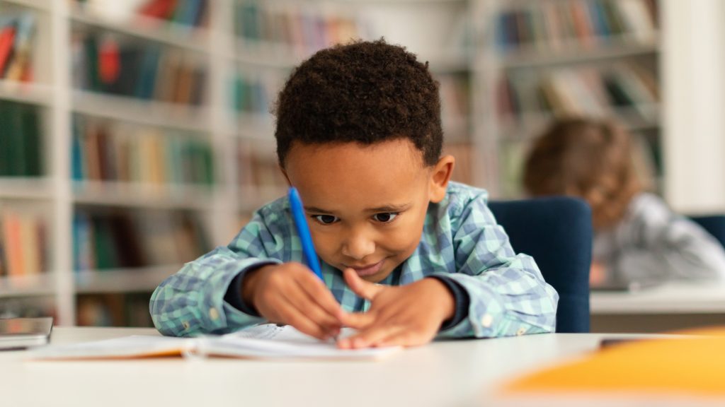 Concentrated black schoolboy sitting at desk in school and learning to write in copybook with pencil